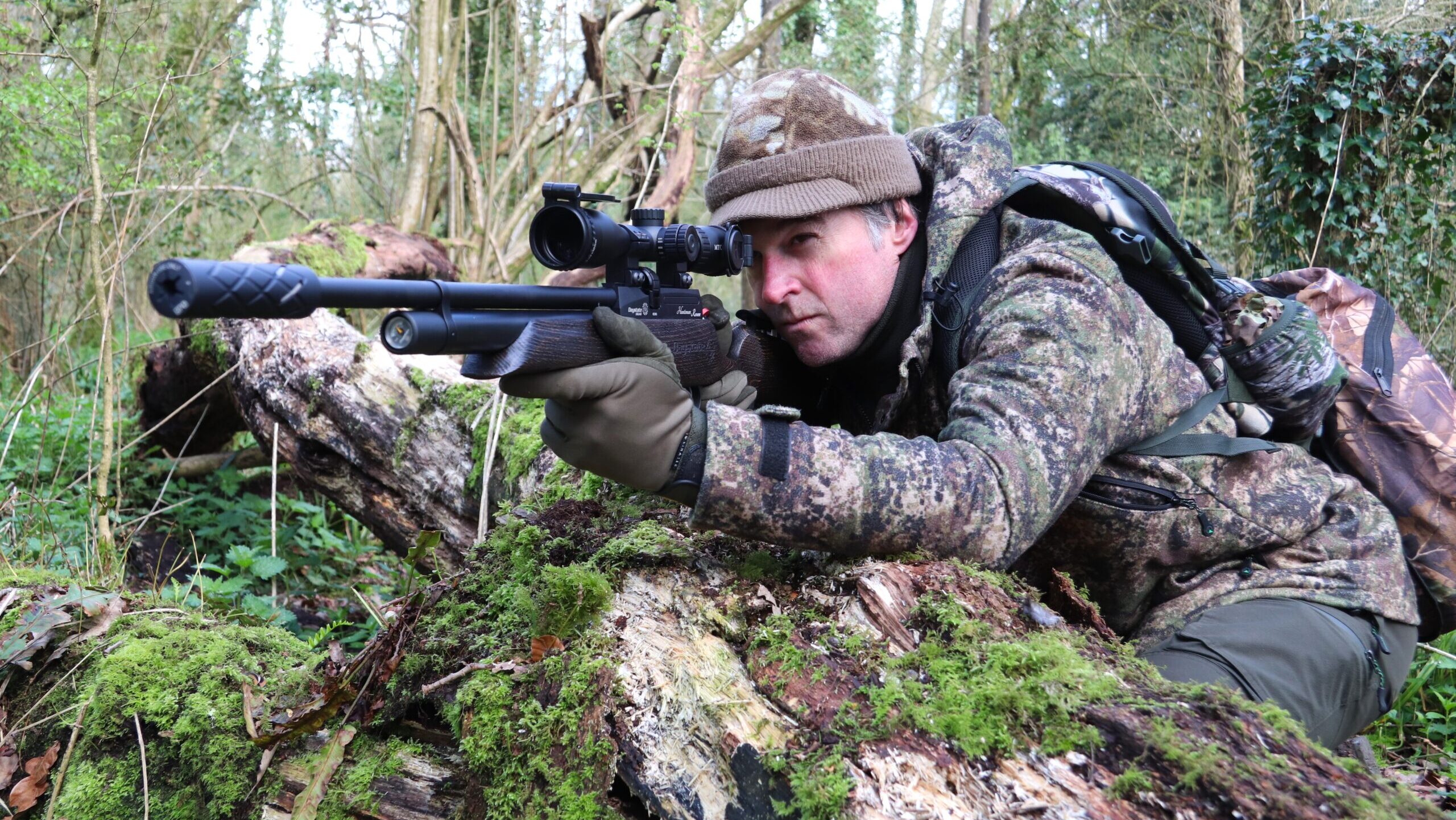 Man in camo leaning on rock as support for air rifle