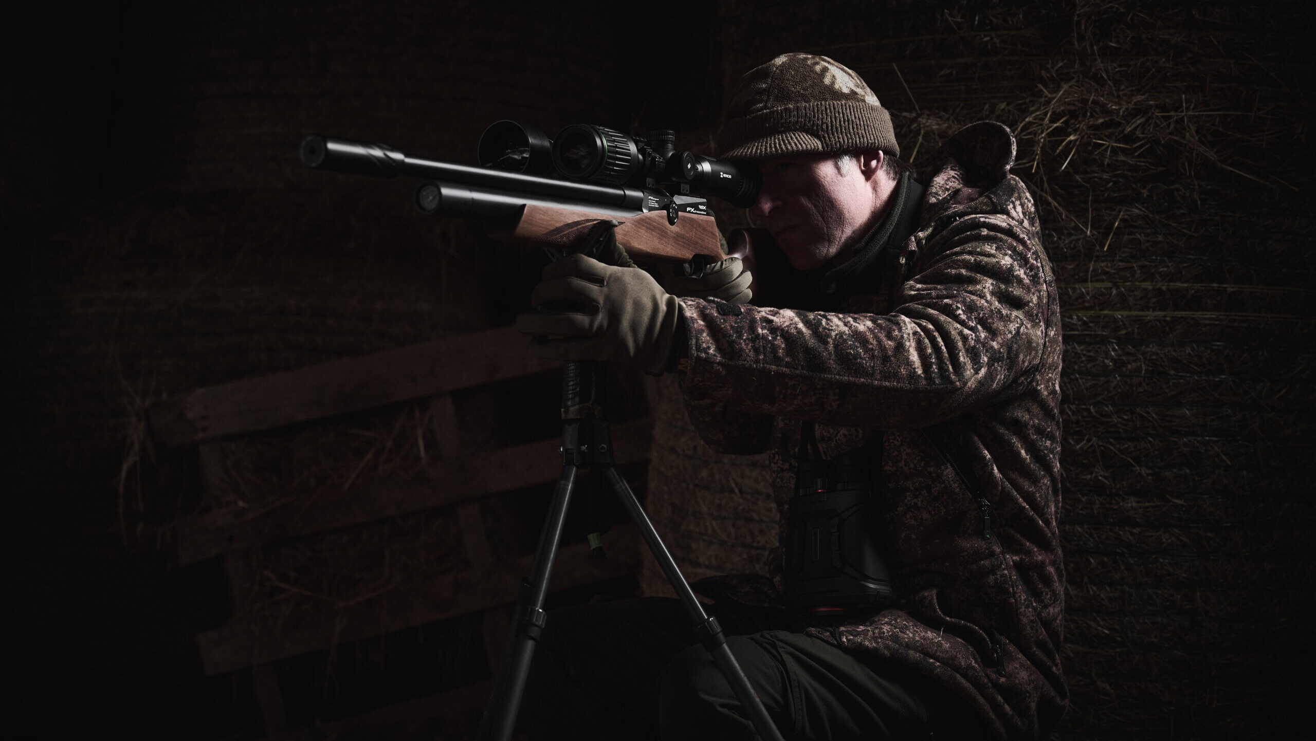 Man in camp crouched on floor with rifle on tripod